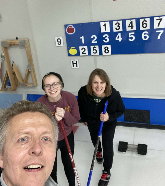 Family of 2 middle-aged adults and 1 twenty-year-old adult holding brooms on a curling rink