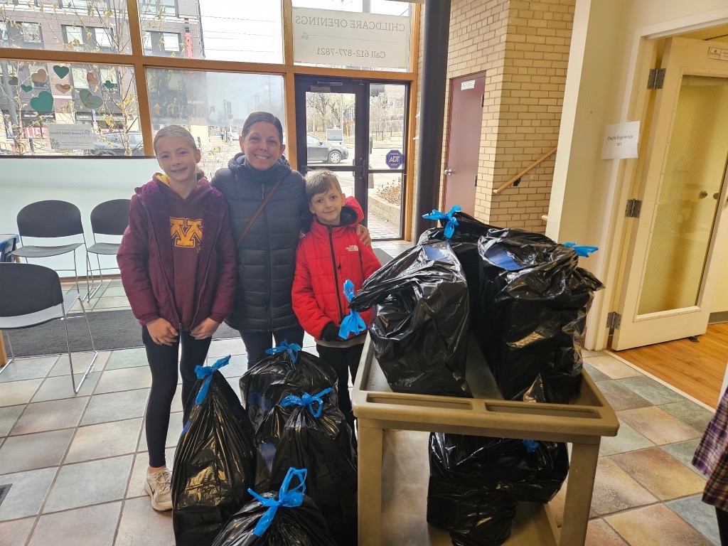 A mom and two children stand with a cart piled with black trash bags filled with gifts.